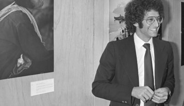 A man in a suit and tie with curly hair and glasses smiles while standing indoors next to a wall displaying black-and-white photographs.
