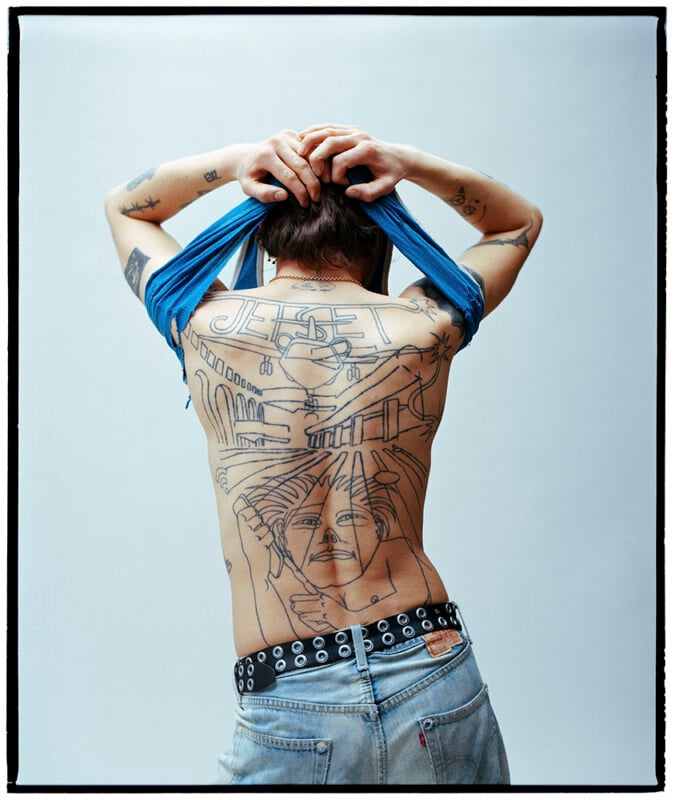 A person with their back to the camera lifts their blue shirt, revealing a large, detailed back tattoo featuring a portrait, architectural elements, rays of light, and the word "JESUS" at the top.