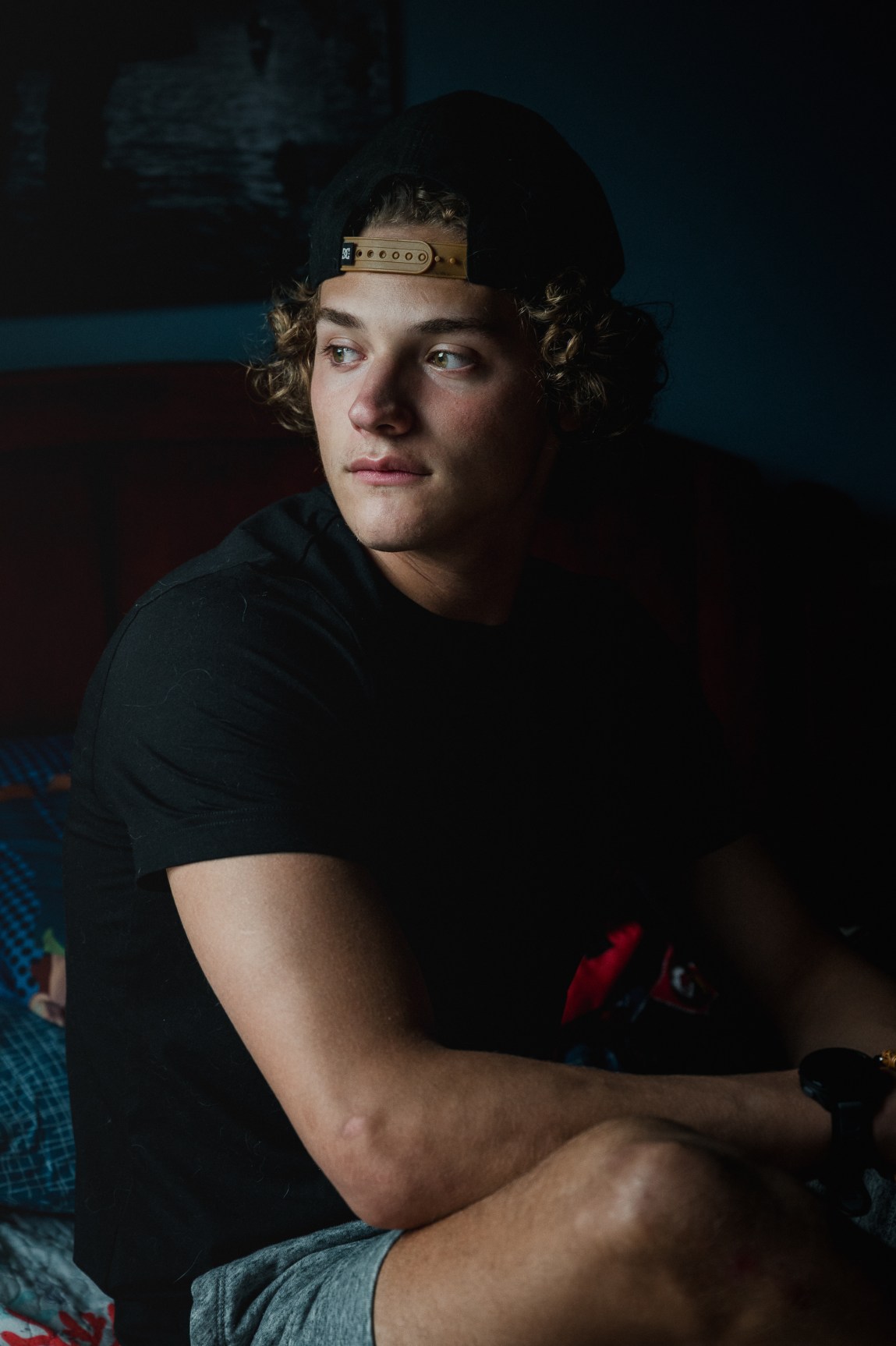 A young man with curly hair and a backward baseball hat gazes off to the distance.