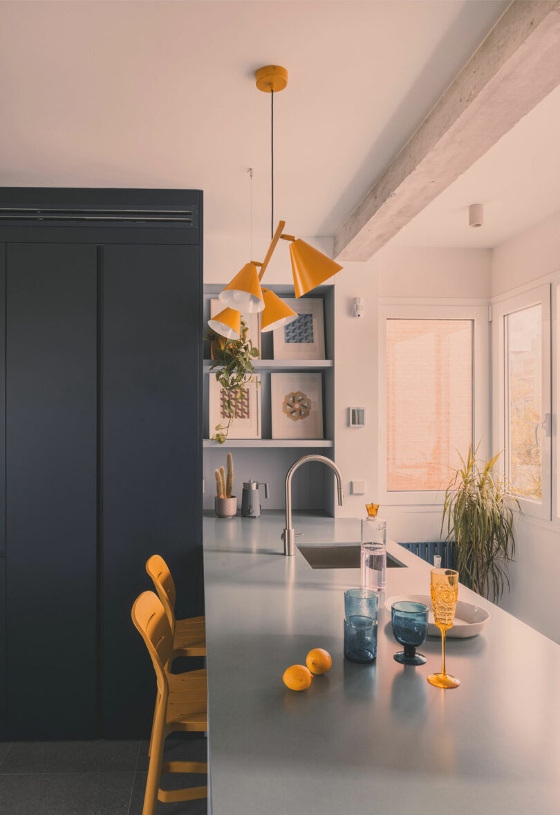 Modern kitchen with a gray countertop, yellow chairs, pendant lights, glasses, lemons, and plants by a window, featuring dark cabinets and minimalist decor.
