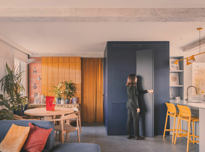 A woman stands by a dark blue cabinet in a modern, colorful apartment with yellow and orange accents, plants, and contemporary furniture.
