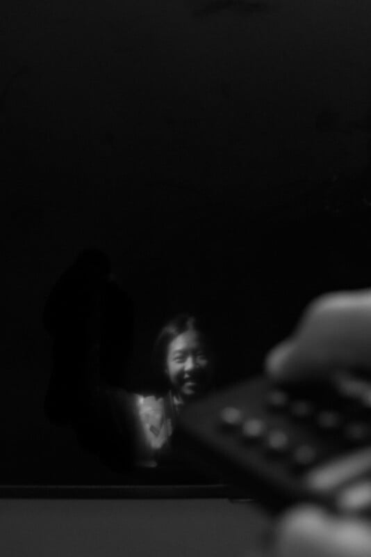 A person’s reflection is visible in a dark screen, smiling faintly. In the foreground, a hand holds a remote control pointed toward the screen. The overall mood is dim and shadowy.