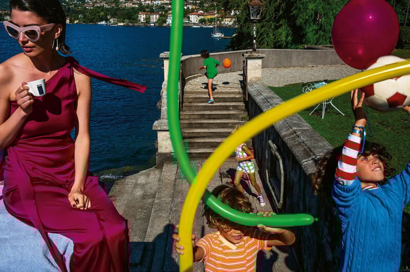 A woman in a magenta dress drinks coffee by a lakeside, while children play with balloons, a soccer ball, and run near stone steps under bright sunlight. A boat floats on the lake in the background.