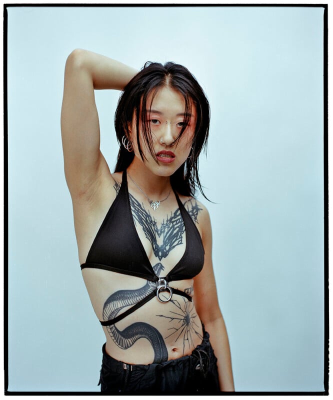 A person with long dark hair wears a black bikini top and black pants, revealing bold, intricate black tattoos on their chest, torso, and arm. They pose with one arm raised behind their head against a plain light background.