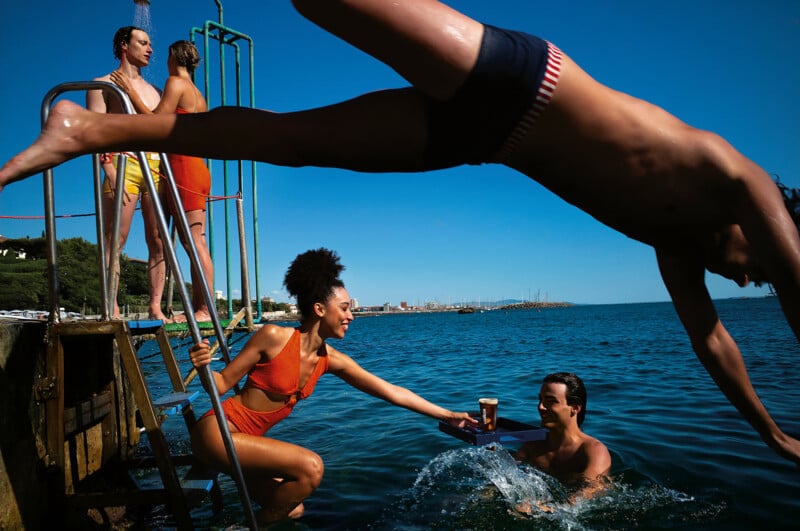 Four people in swimsuits stand on a dock ladder above the water, while one person dives over the group and another in the water receives a drink from a woman on the ladder. The weather is sunny with a clear blue sky.