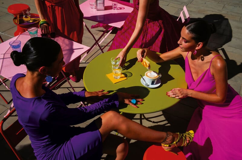 Four women in vibrant dresses sit around a green table outdoors, with drinks and a cup of coffee. Bright sunlight casts dramatic shadows across the colorful scene and pink chairs. One woman dips a pastry into her coffee.