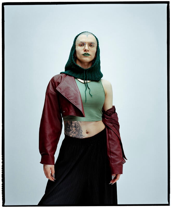 A person stands facing forward, wearing a green knit hood, green crop top, one-sleeved red leather jacket, black skirt, and glasses. They have green lipstick and a large tattoo on their exposed midriff.
