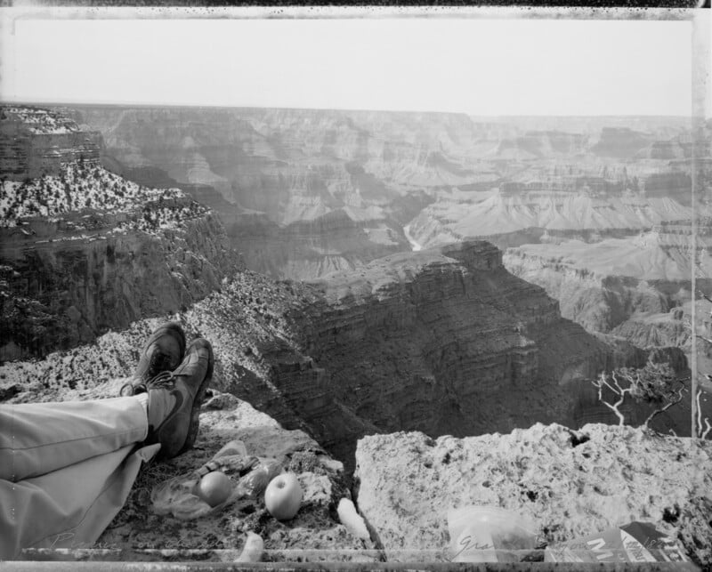 A person’s legs and feet rest on a rocky edge overlooking the vast, layered cliffs of the Grand Canyon; apples, snacks, and a drink are set on the ground beside them. The scene is in black and white.