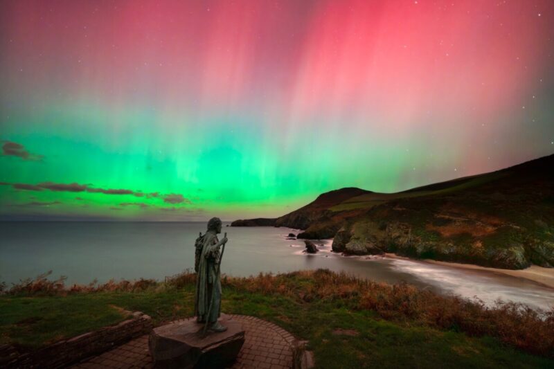 Dramatic coastline with a statue of a man in foreground overlooking the shore and colourful aurora on the sky.