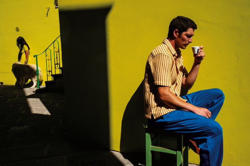 A man in a yellow-striped shirt and blue pants sits on a green stool, drinking from a cup, in front of a bright yellow wall. In the background, a woman walks a large white dog up some stairs in the sunlight.