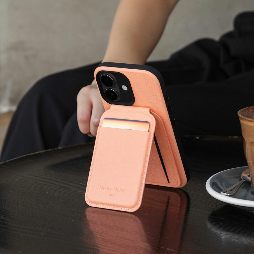A person holding a smartphone with a peach-colored wallet stand case on a table next to a cup of coffee.