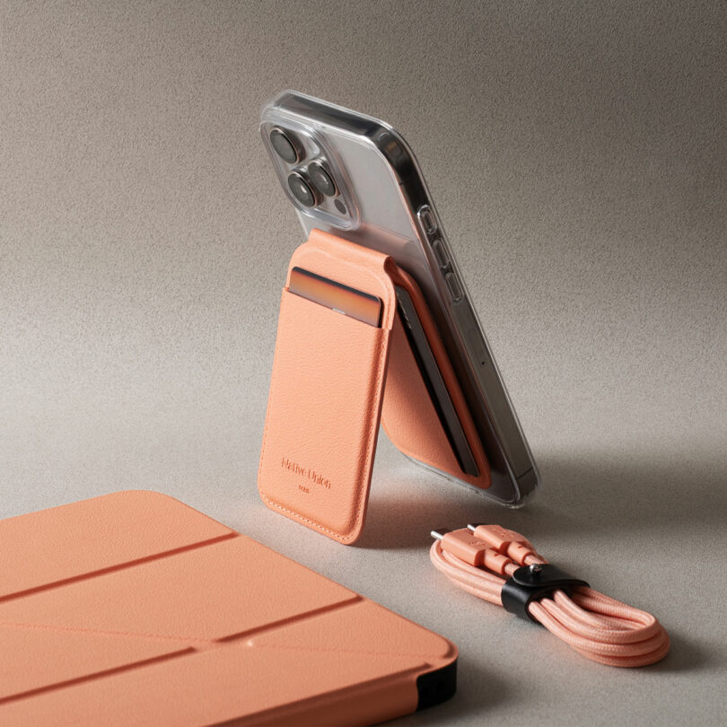 A smartphone with a peach-colored stand and card holder is propped up on a table. Nearby are a matching tablet cover and a coiled charging cable.