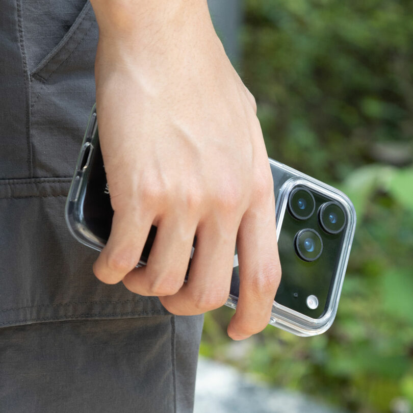 A person holding a smartphone with three rear cameras in their right hand, standing outdoors near greenery.