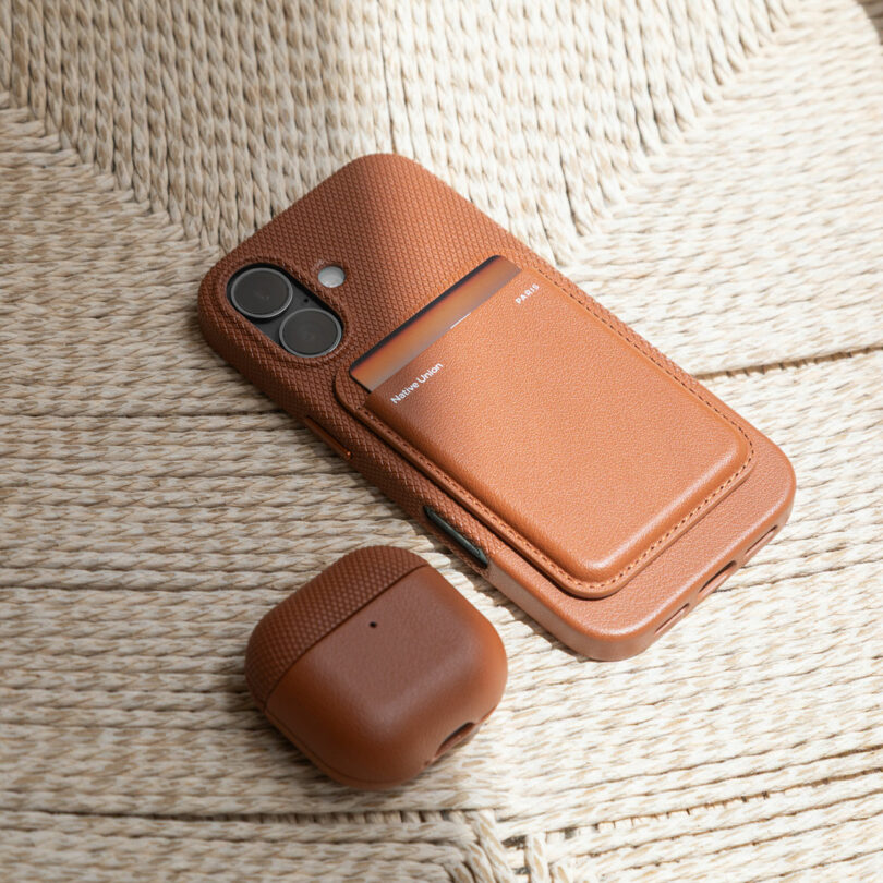 A brown textured phone case with a card holder and a matching wireless earbud case are placed on a woven beige surface.