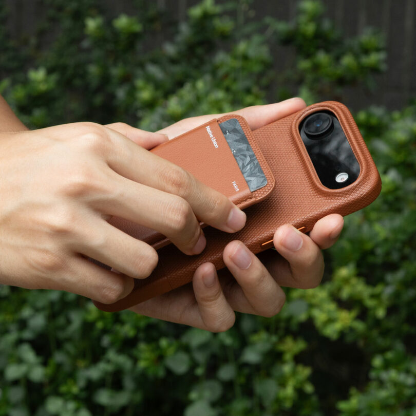 A person holds a phone with a brown leather case and slides a card out of a card slot on the back. Green foliage is visible in the background.