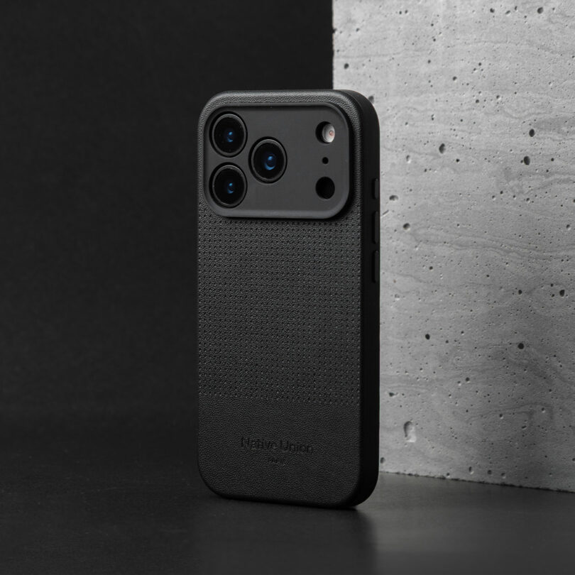 A black smartphone with a textured protective case stands upright against a gray concrete surface and a black background.