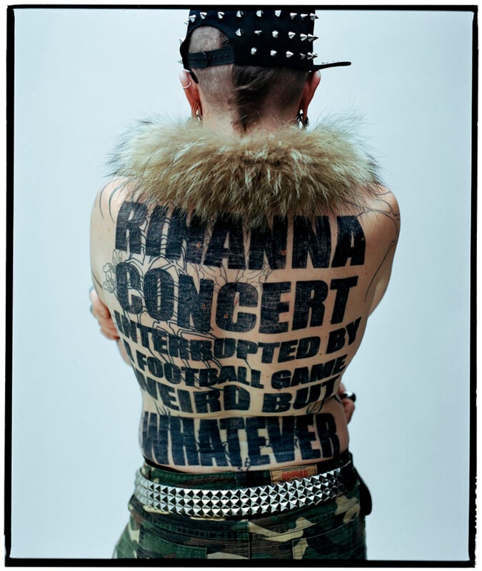 A person with a spiked hat, fur collar, and studded belt shows their back tattoo, which reads: “RIHANNA CONCERT INTERRUPTED BY A FOOTBALL GAME WEIRD BUT WHATEVER.”.