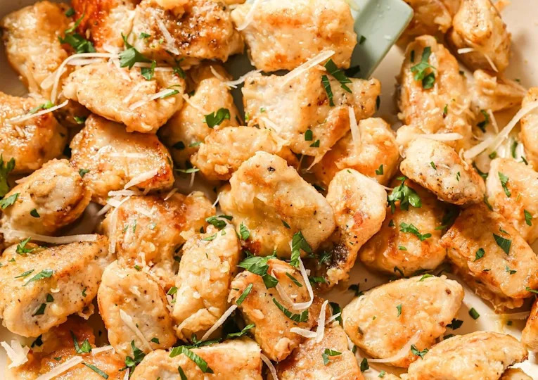 Golden, bite-sized pieces of cooked chicken garnished with chopped parsley and shredded cheese, served on a white plate. The chicken looks crispy and flavorful.