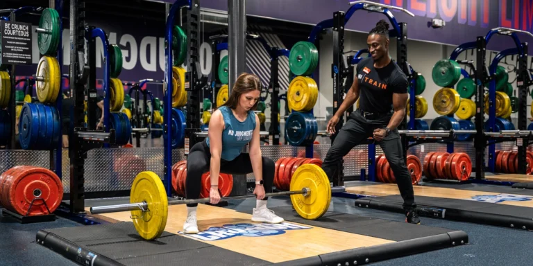 woman picks a barbell up off the ground next to her personal trainer
