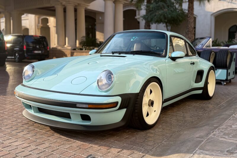 A light blue vintage sports car with white wheels is parked on a brick driveway in front of a building with columns and palm trees, reminiscent of the models featured in "20 Cars That Focused on Analog Engagement in 2025.