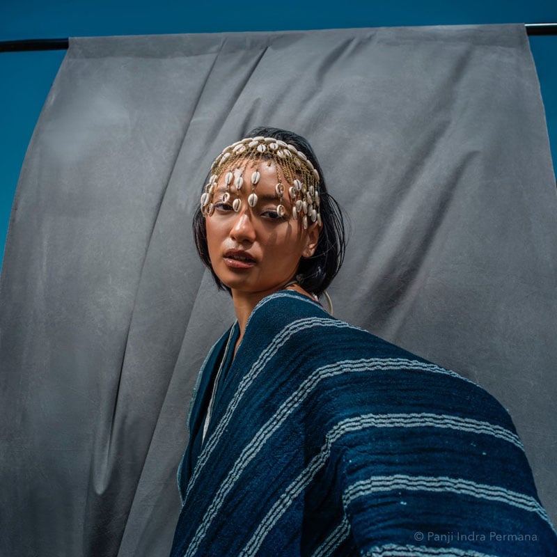 A woman wearing a blue striped shawl and a headpiece decorated with hanging shells poses in front of a blue and gray fabric background.