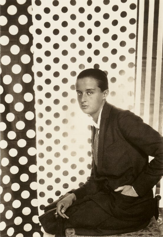 A person in a suit sits sideways on a stool, looking at the camera, with a background of large polka dots and vertical stripes in black and white.