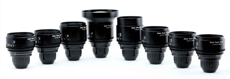 Nine black Zeiss Super Speed camera lenses of varying sizes are lined up in a row against a white background. Each lens has white markings and is positioned upright, showing their different focal lengths.