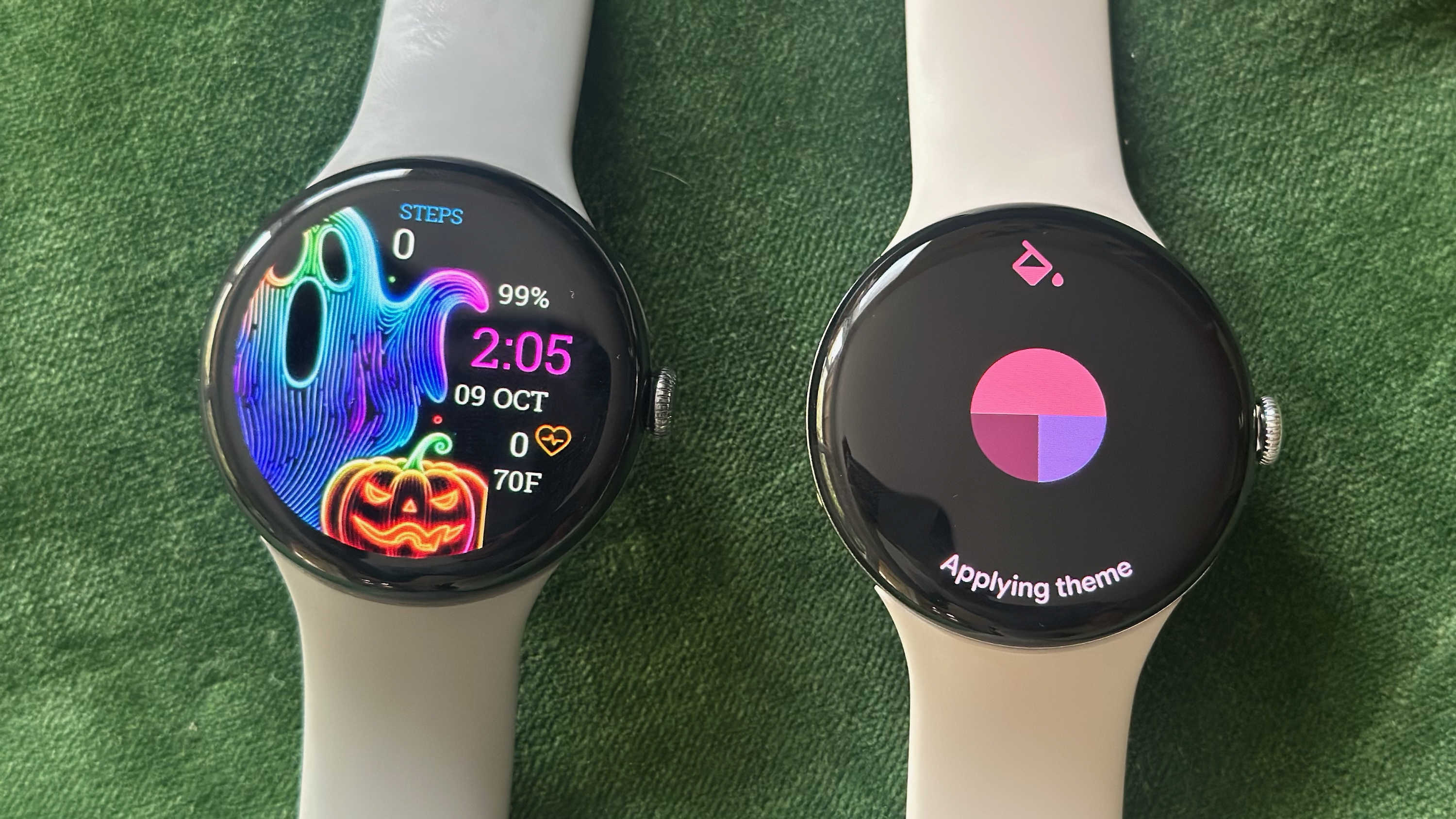 The Pixel Watch 3 (left) and Pixel Watch 4 (right) sitting on a green pillow. The Watch 3 shows a custom Halloween-themed watch face; the Watch 4 shows an "Applying theme" screen pulling red, maroon, and purple colors from a watch face.