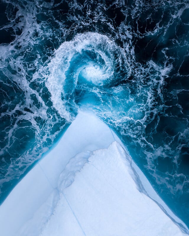 Aerial view of an iceberg with turquoise water swirling in a spiral pattern around its tip, creating dramatic, frothy waves against the deep blue sea.