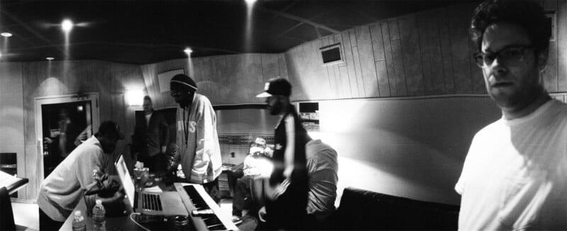 A group of people are gathered in a dimly lit music studio, some standing and some sitting around keyboards and laptops; one person in glasses stands in the foreground, looking toward the camera.