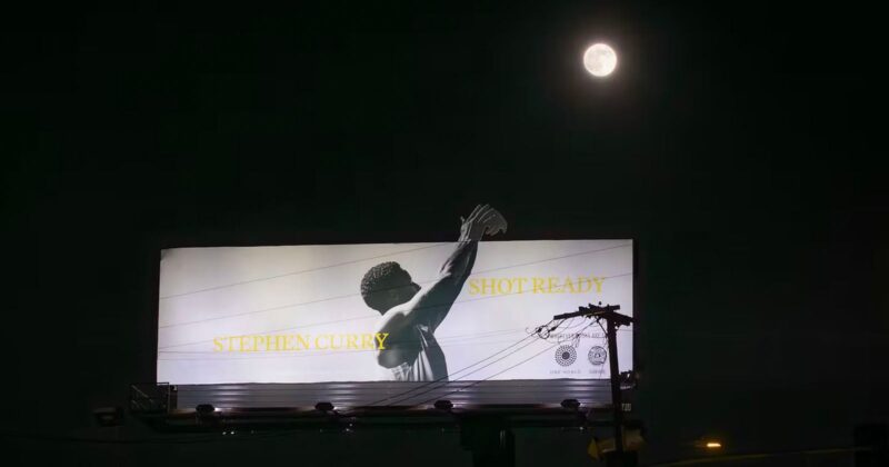 A billboard at night shows a black-and-white image of a basketball player shooting, with the words "STEPHEN CURRY" and "SHOT READY." A full moon glows above the billboard in the dark sky.