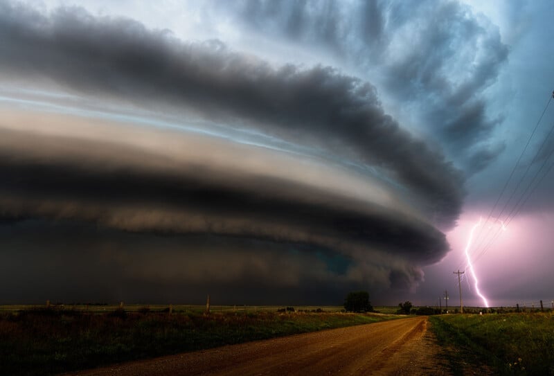 A massive, dark, layered supercell cloud dominates the sky over a rural dirt road, while a bright lightning bolt strikes near power lines in the distance.