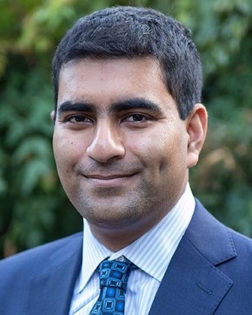 Smiling man with short hair wearing a blue suit and checkered tie.