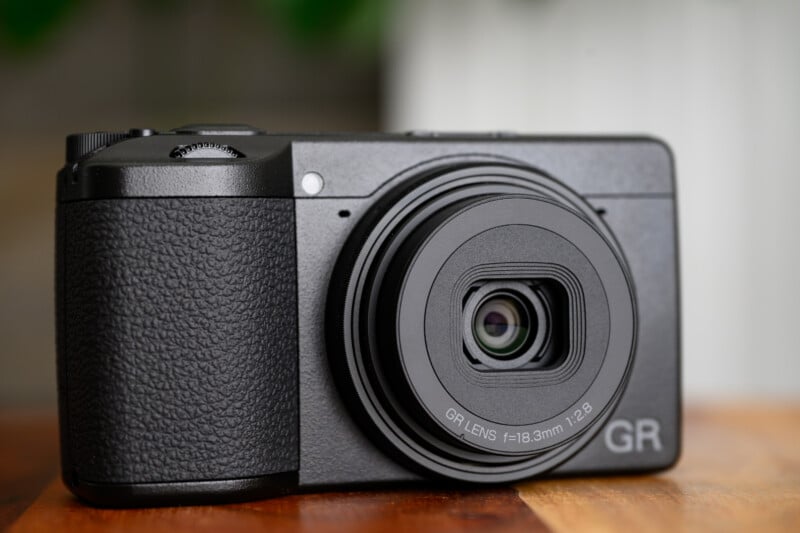 A close-up view of a black compact digital camera with a textured grip, extended lens, and the letters "GR" on the bottom right, placed on a wooden surface.