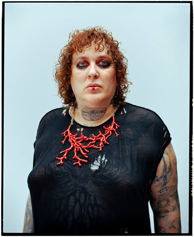 A person with curly, reddish hair and heavy eye makeup stands against a light background. They wear a sheer black top, a bold red coral-shaped necklace, and have tattoos visible on their neck and arms.