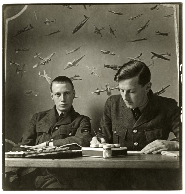 Two men in military uniforms sit at a desk. One looks toward the camera while the other writes. The wall behind them is covered with illustrations of airplanes in flight.