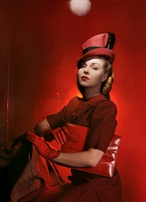 A woman in vintage red attire, including a hat, gloves, and clutch purse, poses against a vivid red background, exuding elegance and poise.