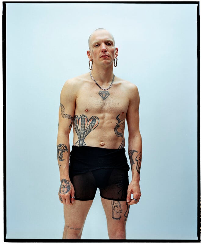 A shirtless person with tattoos, large hoop earrings, a chain necklace, and black sheer shorts stands against a plain light blue background, looking directly at the camera.