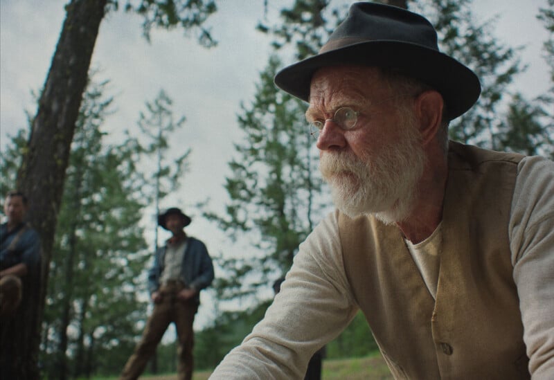 An older man with a white beard, wearing glasses, a black hat, and a beige vest, sits outdoors in a forest, looking thoughtful. Two men stand in the blurred background among tall trees.