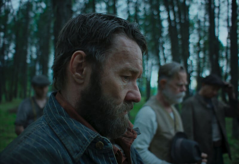 A man with a beard and wearing a denim jacket looks downward pensively in a forest, with three other men standing behind him, blurred in the background. The scene has a somber, muted tone.
