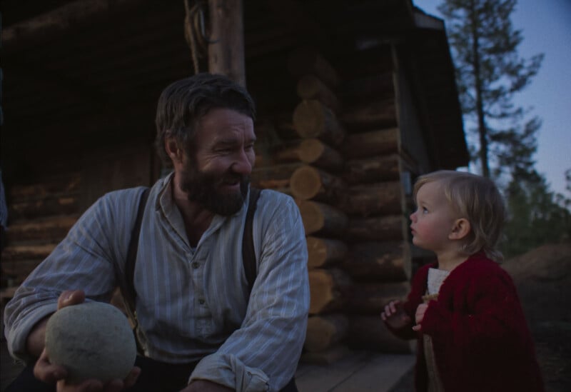 A bearded man in a striped shirt and suspenders smiles at a young child in a red sweater, holding a ball, outside a rustic log cabin at dusk. Trees are visible in the background.