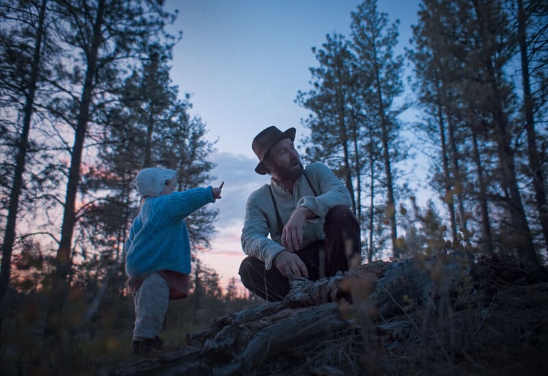 A bearded man in a hat sits on a log in a forest at dusk while a small child in a blue sweater and white hat stands nearby, pointing toward the distance among tall trees.