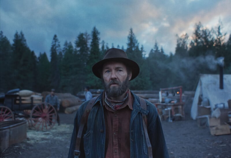 A bearded man wearing a hat and denim jacket stands in a rustic outdoor camp with trees, wagons, and tents in the background under a cloudy sky.