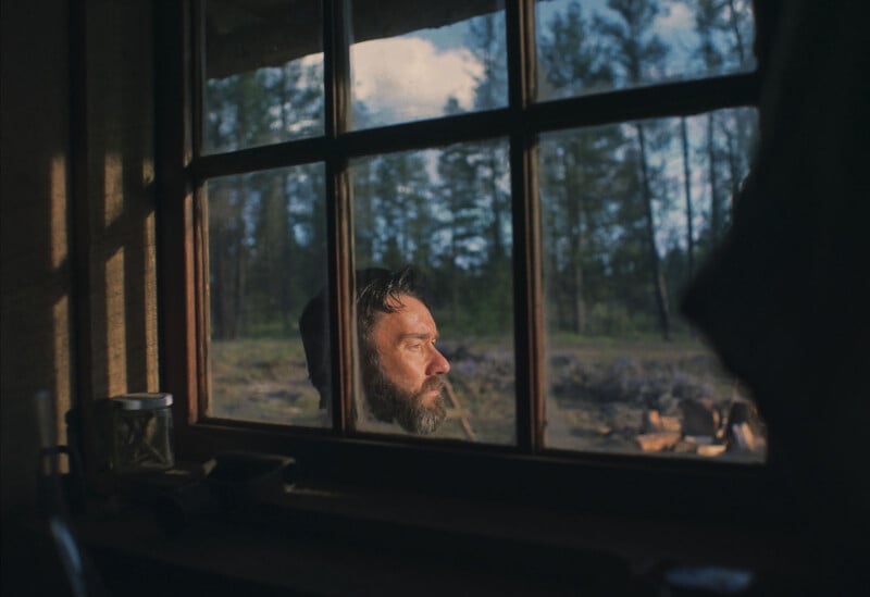 A bearded man is seen through a window, looking pensively into the distance. He is outdoors with a forest and blue sky in the background, while the interior is dimly lit and partially visible.