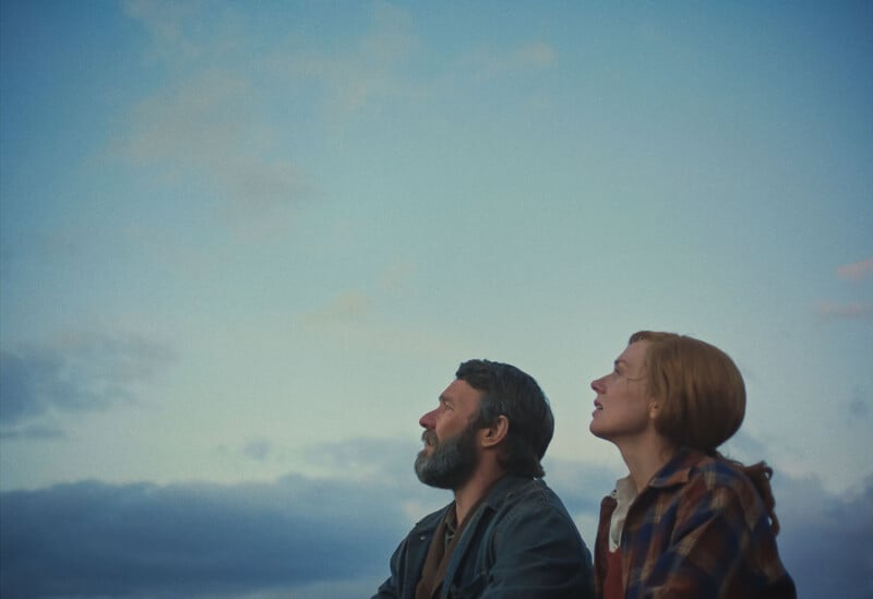 A bearded man and a woman with red hair sit outdoors, looking up at the sky with thoughtful expressions, against a background of blue sky and clouds at dusk.
