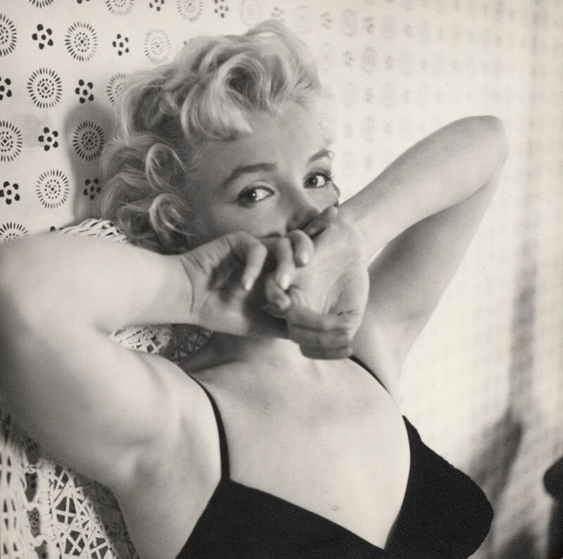 A woman with short, curly blonde hair in a black top sits on a patterned chair, raising her arms and partially covering her face with her hands. The photo is black and white, creating a vintage feel.
