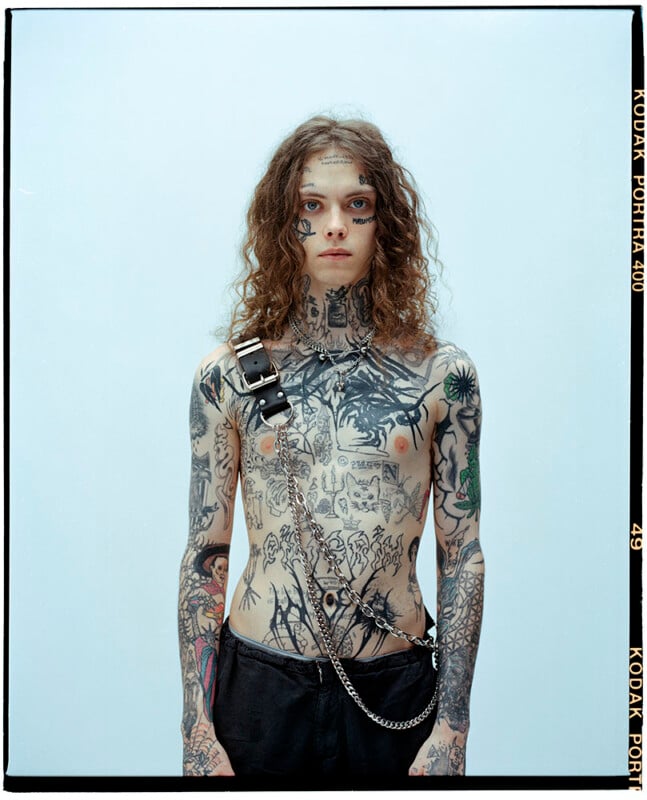 A person with long curly hair stands against a light background, shirtless and wearing black pants. Their upper body and arms are covered in various tattoos, and they wear a harness with a chain across their chest.