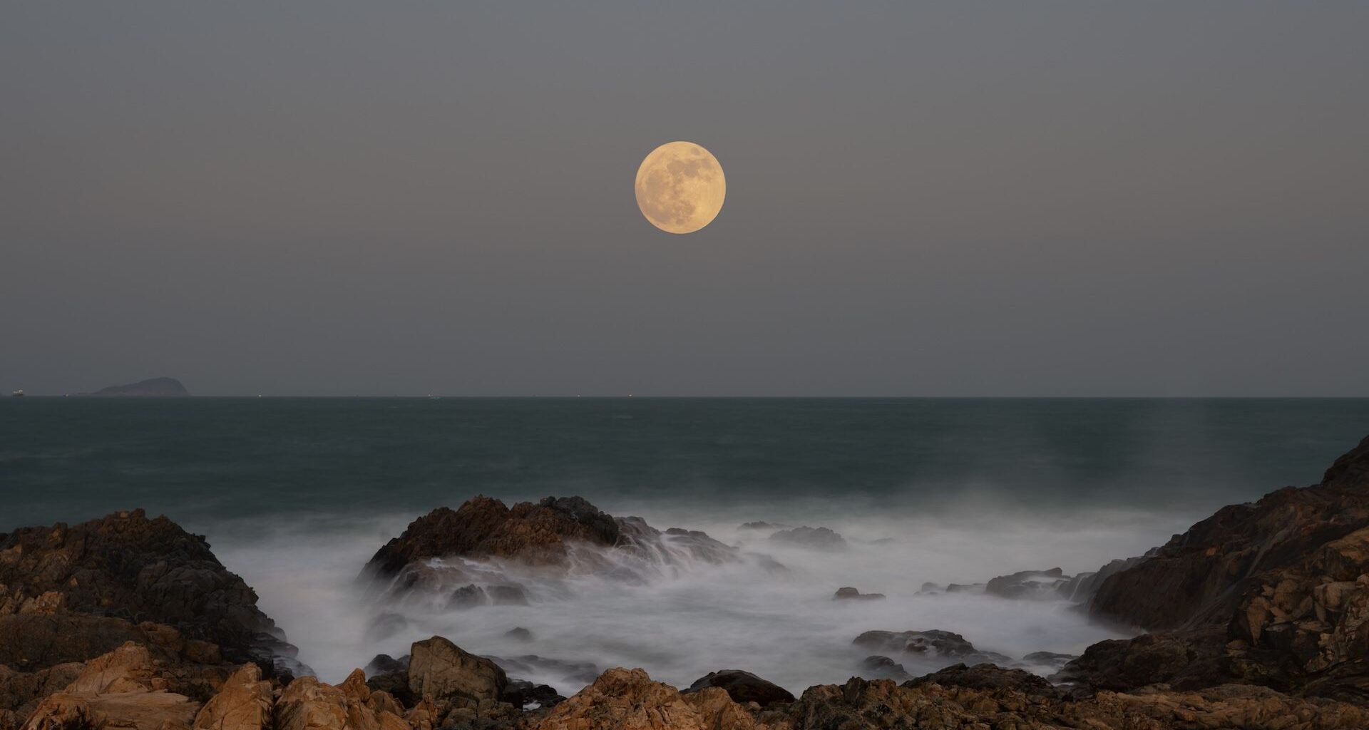 Photo of a full moon over the ocean, taken from rocky shore. Waves can be seen crashing over the rocks in the foreground/bottom of the image.