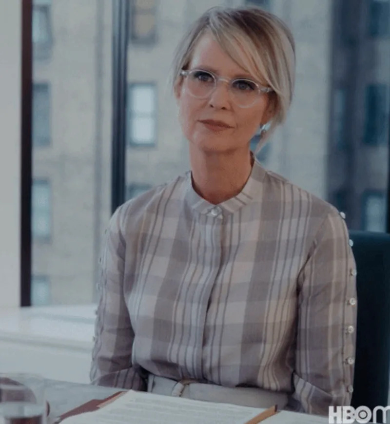 Person with short hair and glasses wearing a plaid blouse, sitting at a desk with an open book, appears thoughtful. Office window in the background