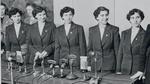 Getty Images the five sisters, wearing suits, lined up in front of microphones on a table, and smiling